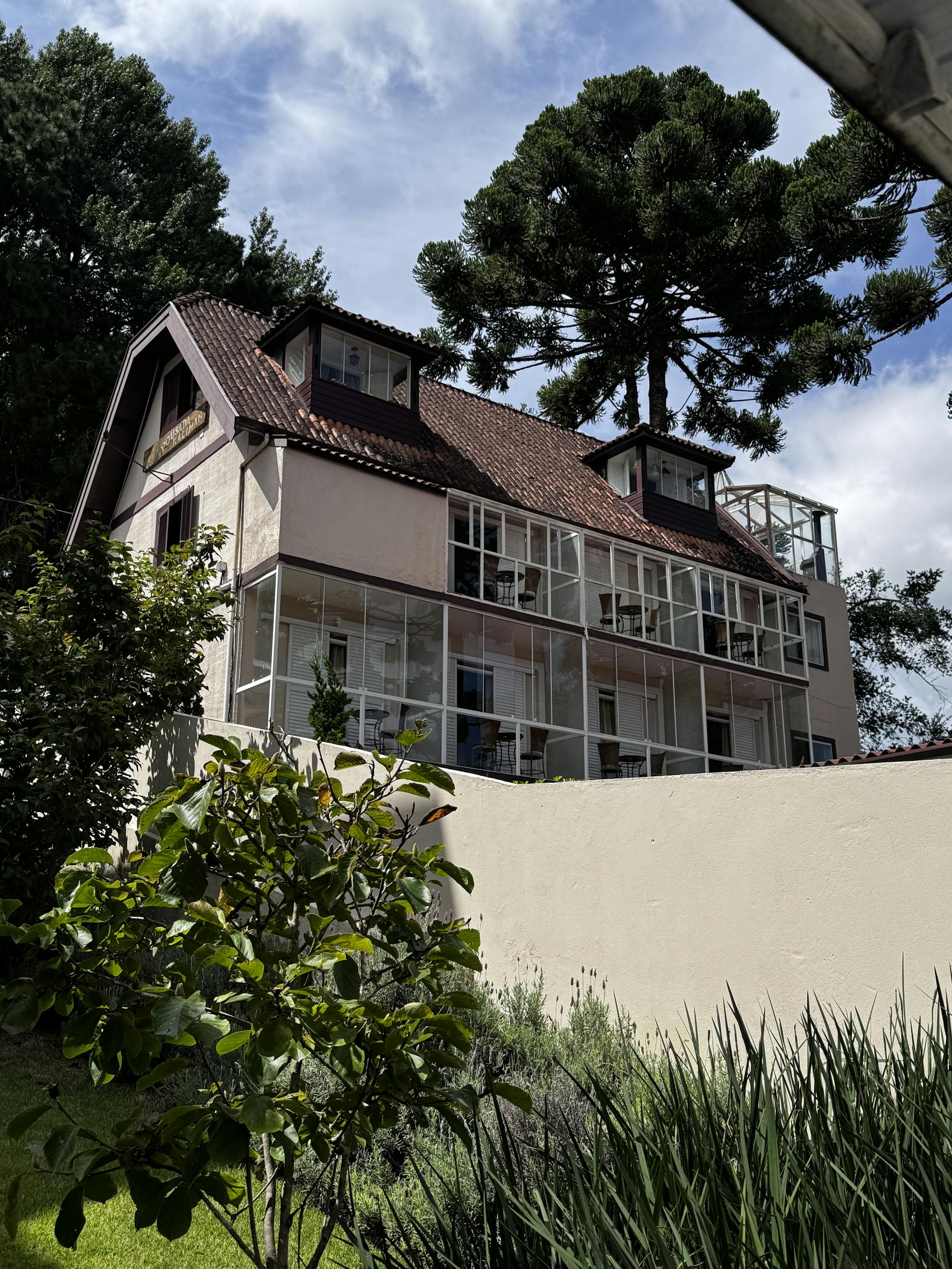 A multi-story house with a brown tiled roof and glass-enclosed balconies set against a backdrop of pine trees.